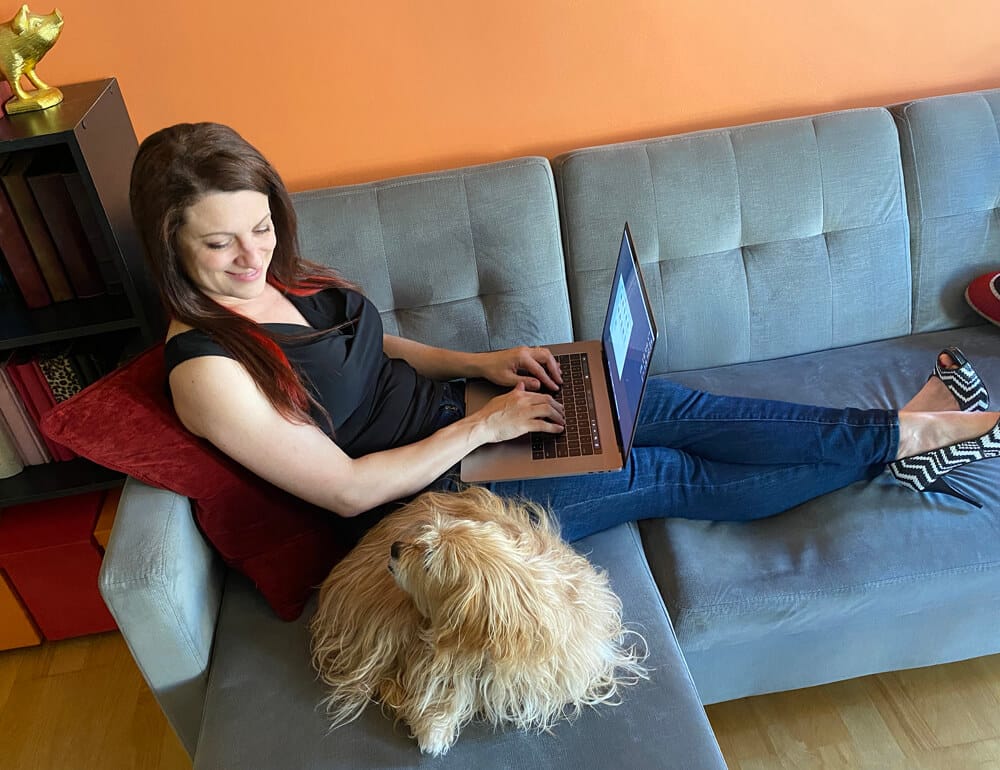 Susy working on laptop on couch with dog by her side